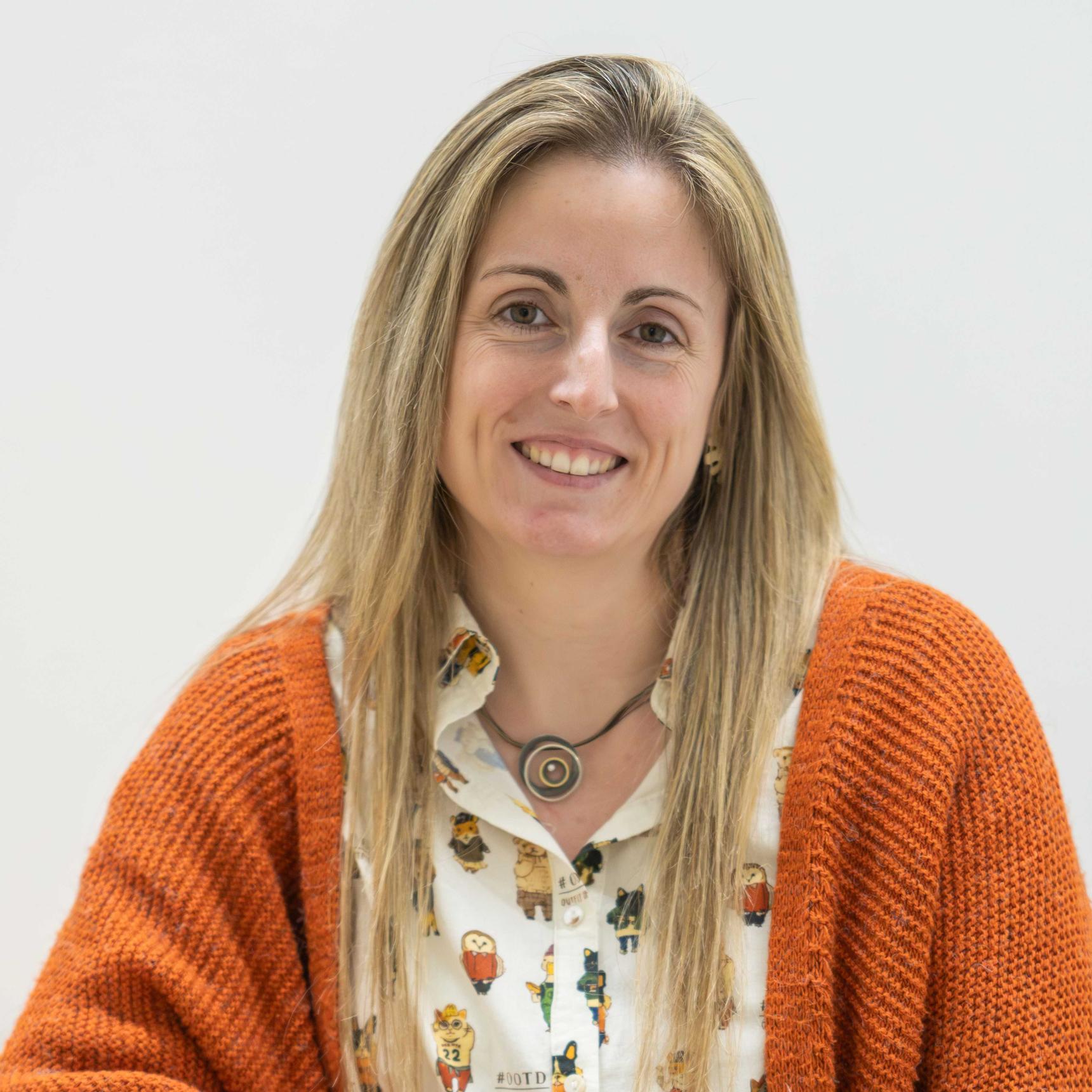 Femme avec des longs cheveux blondes. Elle porte un collier, une veste orange et une chemise à motifs.